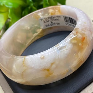 May include: A white agate bangle with brown and orange swirls. The bangle has a label that reads "TT Nghiên Cút Kim Cương & Ba Quy TN VDO199".