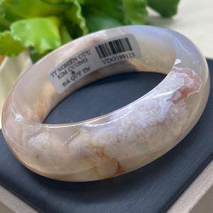 May include: A translucent, oval-shaped bangle bracelet with a marbled pattern of white, beige, and reddish-brown hues. The bracelet has a smooth, polished surface and is set against a dark background. The text on the bracelet is partially visible.