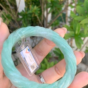 May include: A light green jade bangle bracelet with a twisted rope design. The bracelet is held in a hand, with a tag attached. The tag reads "NATURAL FEI CUI JADE". The background is blurred green foliage.