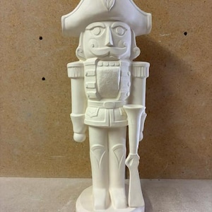 May include: A white, unpainted ceramic nutcracker figurine standing on a circular base. The figure wears a tall hat with a feather detail, a military-style jacket, and holds a long rifle. A ruler shows the height is approximately 16 inches.