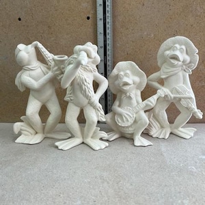 May include: Four unpainted ceramic frog figurines in various poses, each playing a musical instrument. One frog plays a saxophone, another a clarinet, a third a banjo, and the last a violin. The frogs are wearing hats and scarves.
