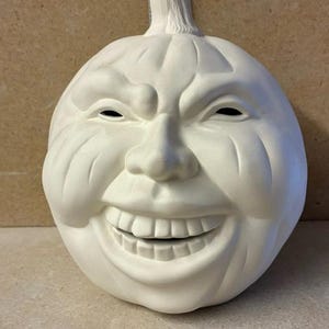 May include: A white ceramic pumpkin sculpture with a carved, grinning face featuring prominent teeth and deep-set eyes. A ruler is positioned behind the pumpkin, showing a height measurement of approximately 8 inches from the base to the stem.