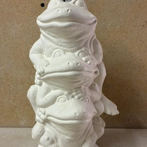 May include: A white ceramic sculpture featuring three stacked frogs. The top frog is positioned above the middle frog, which rests on the bottom frog. A metal ruler is visible behind the sculpture, showing a height of approximately 9.5 inches.
