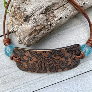 May include: A brown leather bracelet with a copper plate featuring an embossed floral design. The bracelet has two turquoise blue glass beads.