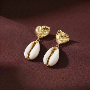 May include: Gold-tone earrings with a seashell design. The earrings have a textured gold top and a white seashell pendant with gold accents. The earrings are displayed on a burgundy background.