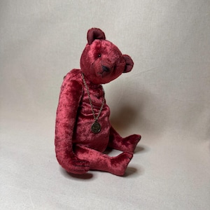 May include: A plush, burgundy teddy bear with a gold chain and a round pendant. The bear is sitting on a white background.