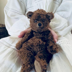 May include: A handmade brown teddy bear with a soft, plush texture. The bear has small, round black eyes, a stitched nose, and a beige paw pad. The bear is a classic toy, perfect for a child or collector.
