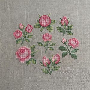 May include: Cross-stitch embroidery of pink roses and buds with green leaves arranged in a circular pattern. The needlework is on a light gray fabric, showcasing the detailed floral design.