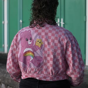 May include: A pink and white chequered jacket featuring a Care Bear design on the back. The design shows a pink Care Bear holding a yellow star, with a rainbow on its belly. The jacket has a cropped style and long sleeves.