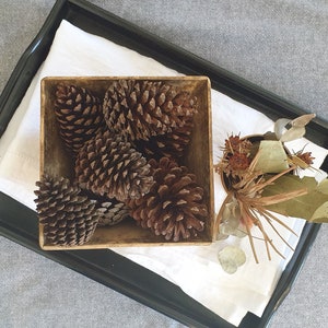 May include: A black tray holds a square wooden bowl filled with brown pine cones, resting on a white cloth. A small arrangement of dried leaves and flowers sits in a small container. The background is a grey fabric.