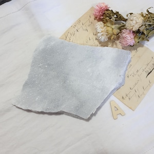 May include: A pale grey, rough-edged stone slab with a sparkling, crystalline surface. The stone sits on white fabric, with vintage paper and dried flowers in the background. A wooden letter "A" is also present.