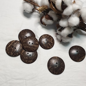 May include: Several dark brown coconut shell buttons, each with two holes, are scattered on a white surface. The buttons have a natural, textured finish. A sprig of cotton with white blooms and brown pods is in the background.