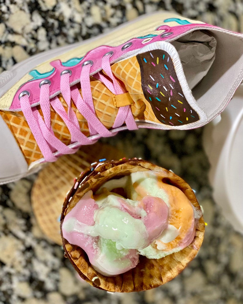 May include: A pair of white high-top sneakers painted to look like an ice cream cone. The sneakers have pink laces and a brown and white waffle cone design. The ice cream is pink, green, and orange.