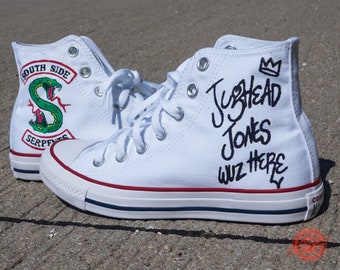 May include: White high-top Converse sneakers with custom hand-painted designs. One shoe features a green snake with the words "South Side Serpents" and the other shoe has the words "Jughead Jones Wuz Here" with a crown.