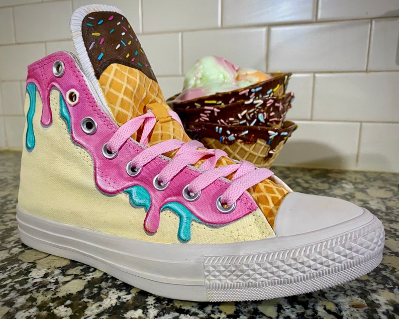 May include: A pair of white high-top sneakers with a yellow canvas base and pink and blue ice cream dripping down the side. The sneakers have a waffle cone pattern on the side and a brown ice cream cone with sprinkles on the top.