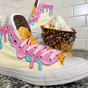 May include: A pair of white high-top sneakers painted to look like ice cream cones. The sneakers have pink, blue, and yellow dripping ice cream designs. One sneaker has a waffle cone design and the other has a chocolate-dipped waffle cone design.