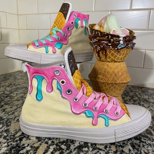 May include: A pair of white high-top sneakers with a pink and blue ice cream drip design. The sneakers have a waffle cone pattern on the side and a brown chocolate sprinkle detail.