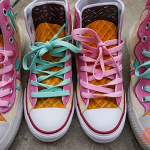 May include: A pair of high-top sneakers painted to look like ice cream cones. The sneakers have a yellow waffle cone design, pink and blue ice cream, and brown chocolate sprinkles.