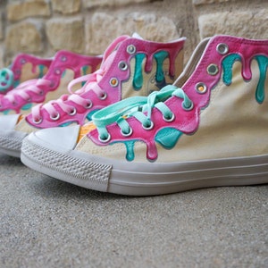 May include: A pair of high-top canvas sneakers painted with pink and teal dripping paint, resembling melted ice cream. The sneakers are on a gray concrete surface.