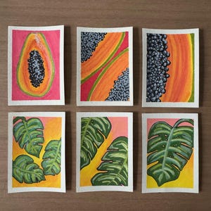 May include: Six watercolor paintings on white paper. The paintings depict papaya slices and monstera leaves. The papaya paintings feature orange and yellow fruit with black seeds. The monstera paintings show green leaves with yellow and pink backgrounds.