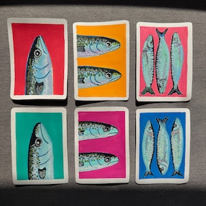 May include: Six colorful, rectangular art pieces featuring painted fish. Each piece has a different background color: red, orange, pink, teal, magenta, and blue. The fish are painted in shades of blue, gray, and white.
