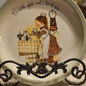 May include: A vintage plate featuring a girl cooking, displayed on a decorative wrought iron stand. The plate has a light green border and the text "A little spice and a lot of nice..." The plate is supported by a decorative stand.