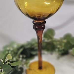 May include: Amber-colored wine glass with a round bowl and a long, slender stem. The base is a flat circle. The glass is made of a transparent material, allowing light to pass through. The stem has a decorative design.