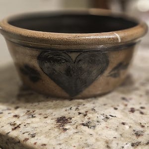 May include: A round, brown ceramic bowl with a black interior. The exterior features a large, hand-painted black heart and smaller black leaf-like designs. The bowl sits on a speckled countertop.