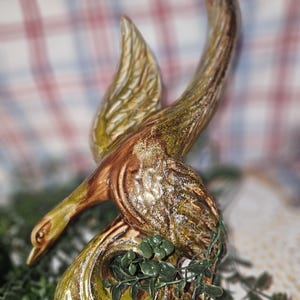 May include: A ceramic bird figurine with a glossy finish in shades of brown, gold, and green. The bird is depicted in a dynamic pose with one wing raised and its head tilted downward. The base features a circular opening and textured details.