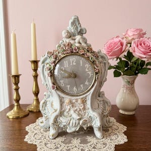 May include: A vintage-style, ornate clock with a light blue and white porcelain finish. The clock features a round face with Roman numerals, cherub accents, and is adorned with pink floral details. Two candles in brass holders and a vase of pink roses are also present.