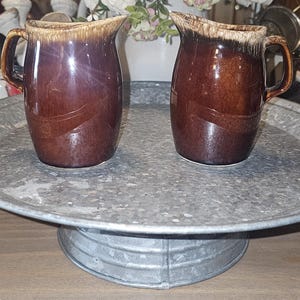 May include: Two matching ceramic pitchers featuring a deep brown glossy glaze with a distinct cream-coloured drip pattern along the top rim. Each pitcher has a curved handle and a pouring spout, showcasing a smooth, reflective finish on the exterior.
