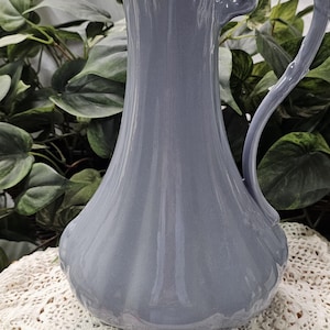 May include: A tall, gray-blue ceramic pitcher with a fluted body and a curved handle. The pitcher has a scalloped rim and a spout for pouring. It is displayed on a white lace tablecloth, with green foliage in the background.