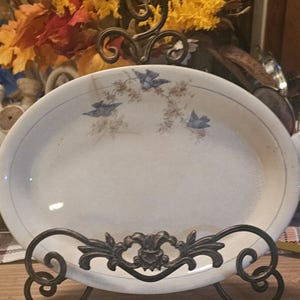 May include: An oval ceramic platter with a cream-colored base, featuring a delicate pattern of three small blue birds perched on thin brown branches. The platter is displayed on a dark brown metal stand with ornate scrollwork details.