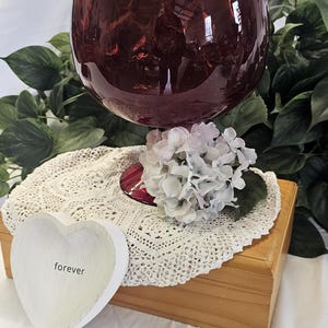 May include: A large, burgundy-colored glass with a wide bowl, resting on a wooden box. A white lace doily and a heart-shaped dish with the word "forever" are in front. A cluster of white and lavender flowers sits near the glass.