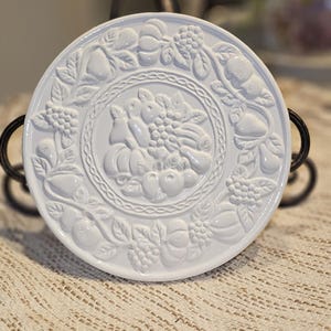 Puede incluir: Plato decorativo de cerámica blanca con diseño en relieve de frutas y verduras. El plato presenta uvas, peras y calabazas. El plato se exhibe sobre un soporte decorativo de metal.