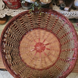 May include: A round woven basket featuring a deep red rim and base accents, with natural tan woven sides and a star-patterned center. The basket has a wide, open top and a structured design made from thin, flexible strips of wood and twisted fiber.