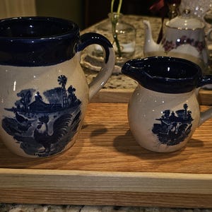 May include: Two ceramic pitchers with blue and white pastoral scenes, including a rooster, sit on a wooden tray. The pitchers have dark blue rims and handles. The tray is rectangular with rounded corners.