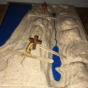 Peut inclure: Diorama religieux représentant une chaîne de montagnes avec une rivière et un lac bleus. Des croix en bois et des étoiles sont placées sur la montagne. Les mots "Bethléem" et "Jérusalem" sont visibles.