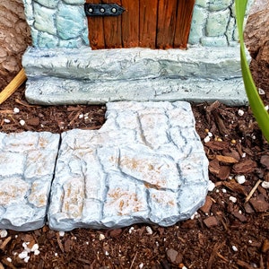 May include: A miniature stone house with a wooden door and a green tree of life design. The house has a stone walkway leading to the door.