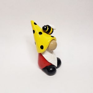 May include: A small gnome figurine with a yellow polka dot hat and a bee on top. The gnome is wearing a red shirt and black shoes.