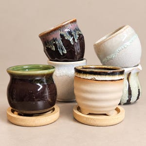 May include: A collection of five small ceramic pots with varied glaze finishes, including dark brown, cream, and textured white, each resting on a circular wooden base. The pots feature rounded shapes and distinct rim details.