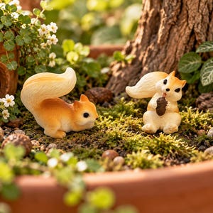 May include: Two small squirrel figurines with cream and orange fur. One squirrel is positioned in a crouching stance, while the other sits upright holding a small brown acorn. Both figures feature bushy tails and dark eyes, displayed among moss.