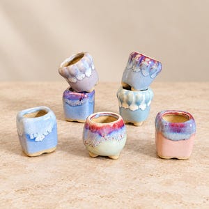 May include: A collection of six small ceramic planters with varied reactive glazes in shades of blue, purple, pink, and cream. Each vessel features a rounded or square shape, a smooth interior, and a distinct drip-style glaze pattern.