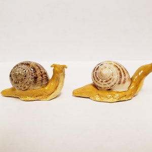 May include: Two ceramic snail figurines. The snails have tan bodies and brown and tan striped shells. The figurines are on a white surface. The snails are facing opposite directions.