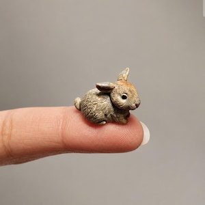 May include: A miniature figurine of a brown and tan rabbit with textured fur, pointed ears, and a small tail. The rabbit is depicted in a seated position with its front paws tucked underneath its body, showcasing detailed facial features.