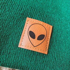 May include: Green knitted beanie with a brown leather patch featuring a black alien face.