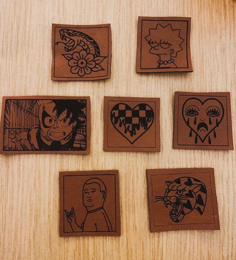 May include: Set of 7 brown leather patches with black ink designs. The designs include a snake with a flower, a cartoon character with yellow hair, a checkered heart, a crying heart, a cartoon character with a rock and roll hand gesture, and a tiger.