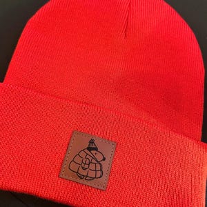 May include: Bright red beanie hat with a folded brim. A square, brown patch with a black cartoon graphic of a bundled-up animal wearing a hat is sewn on the front. The hat is made of a knitted material.