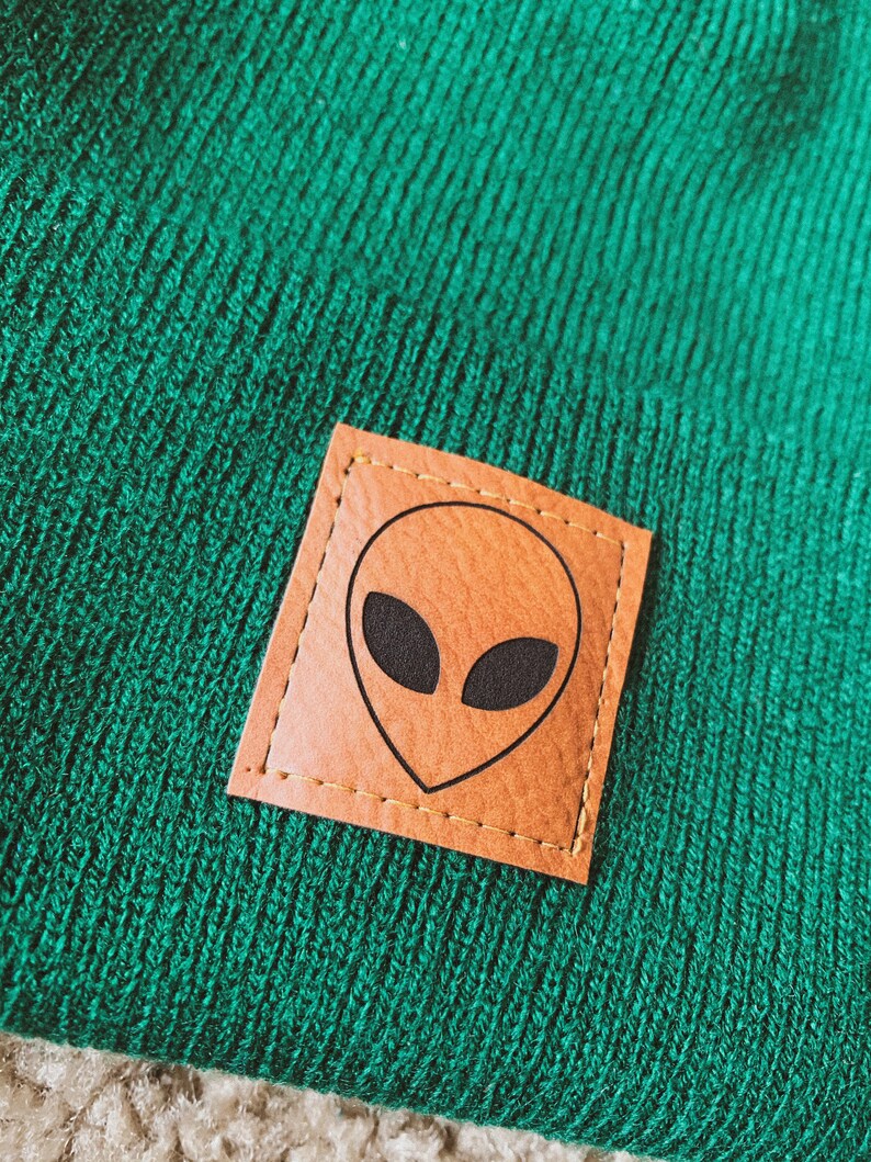May include: Green knitted beanie with a brown leather patch featuring a black alien face.