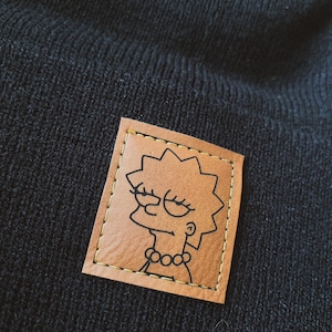 May include: A black beanie with a brown leather patch featuring a cartoon image of a yellow-haired woman with a bored expression. The patch has a gold stitched border.